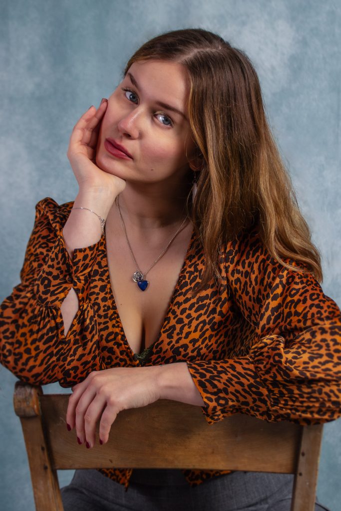 A woman with brown hair leans on the back of a chair, chin resting on hand