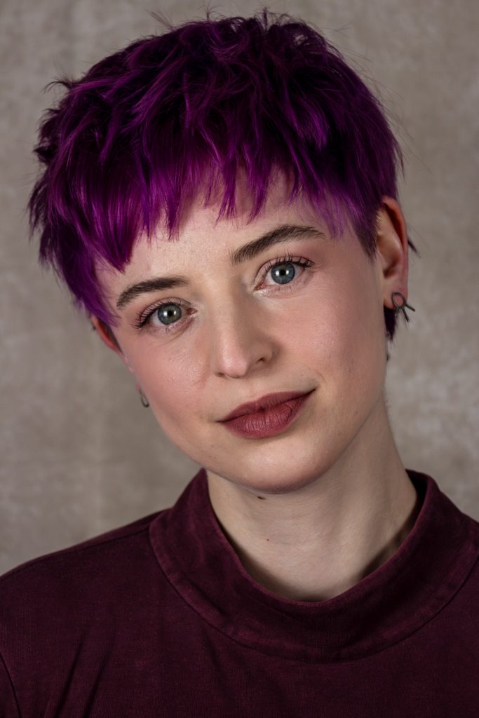 A woman with purple hair looks at the camera
