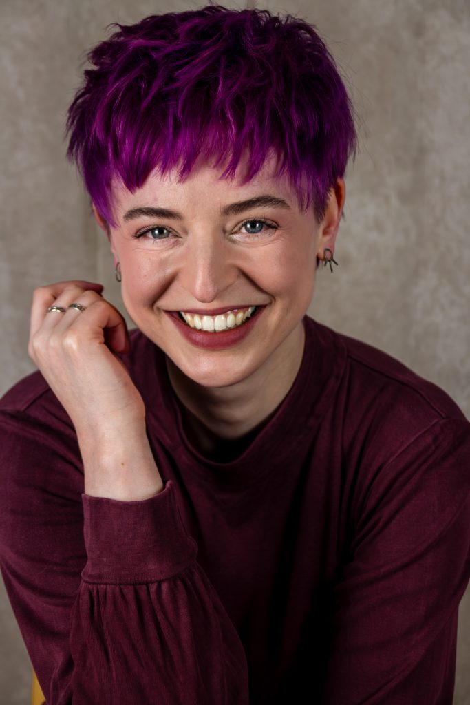 A woman with purple hair is smiling at the camera.
