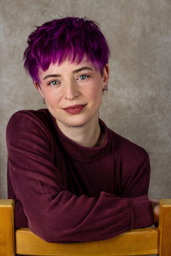 A woman leans on a chair and looks at the camera. She has purple hair.
